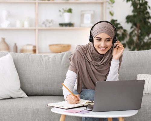 Online Education For Muslim Women. Happy Arabic Girl In Headscarf And Headset Studying With Laptop At Home, Taking Notes While Watching Webinar